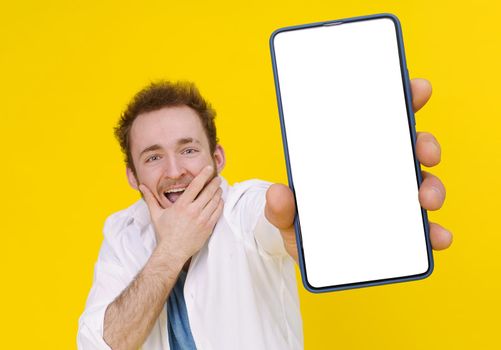 Cant believe, shocked young man with smartphone showing a white empty screen isolated over yellow game, bet, lottery win. Product placement. Mobile app advertisement