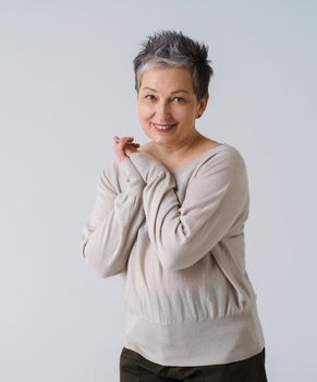 Mature tender grey hair woman in 50s european ethnicity posing looking at camera with hands folded wearing cream blouse isolated on white. Smiling woman healthcare, aging beauty, cosmetics concept