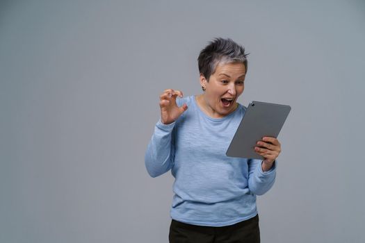 Inspired, shocked mature grey hair businesswoman with digital tablet in hand working, shopping, gaming online. Pretty woman in 50s in blue blouse isolated on white
