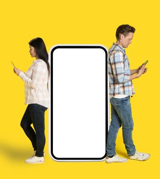 Asian woman and caucasian man standing leaned on huge smartphone with white screen looking at phones in hands, mobile app advertisement isolated on yellow background. Product placement