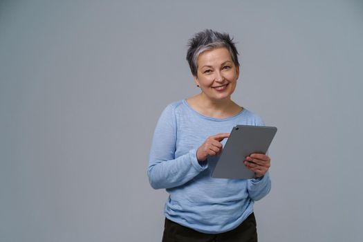 Mature grey haired businesswoman with digital tablet in hands working online. Pretty woman in 50s in blue blouse isolated on white. Older people and technologies