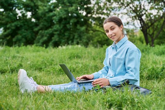 Teenage girl high school student using laptop while sitting on green grass, looking at camera. Adolescence, nature, education, technology concept