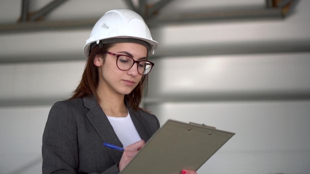 Young woman in a helmet with documents at a construction site. The boss woman in a suit keeps records of documents on architecture. The girl looks around.