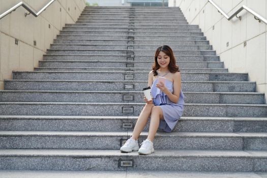 Cheerful young Asian woman holding takeaway coffee cup, using mobile phone