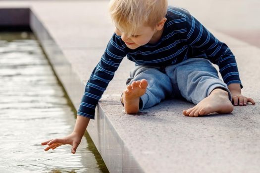A little funny boy climbed on a parapet of a fountain and touch the water.