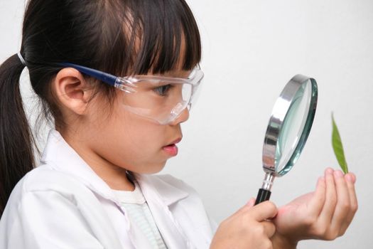 Portrait of a little girl in glasses holding a magnifying glass looking at leaves in researcher or science uniform on white background. Little scientist.
