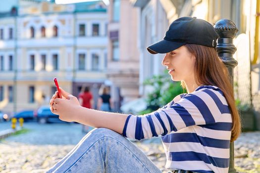 Urban style, fashionable teenage female sitting on the sidewalk using smartphone
