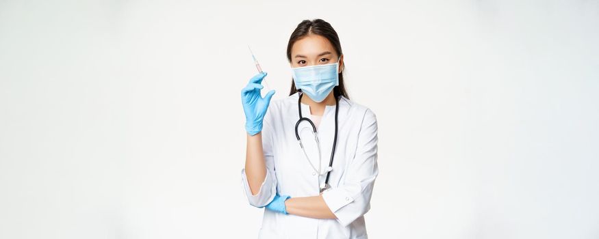 Vaccination and healthcare concept. Asian female doctor, nurse in medical face mask and gloves holding syringe with vaccine, white background