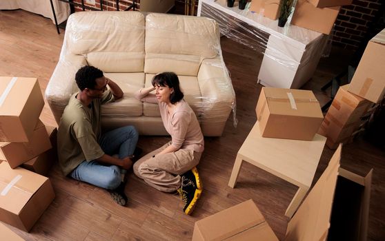 Diverse husband and wife sitting on apartment floor and enjoying relocation