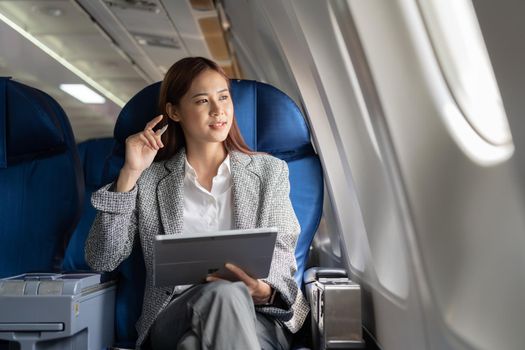 Successful beautiful young asian business woman sits in airplane cabinplane and works on digital tablet with stylus. Flying at first class.
