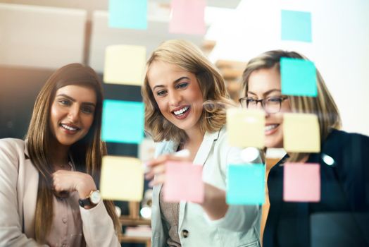 Brainstorming their way to brilliant business. Shot of a group of businesswomen arranging sticky notes on a glass wall in a modern office.
