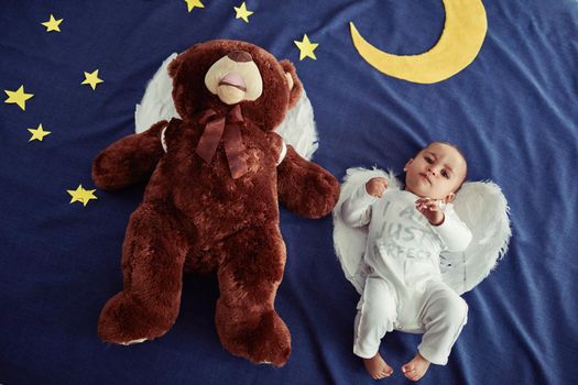 Concept shot of an adorable baby boy and a teddy bear wearing angel wings against an imaginary night time background.