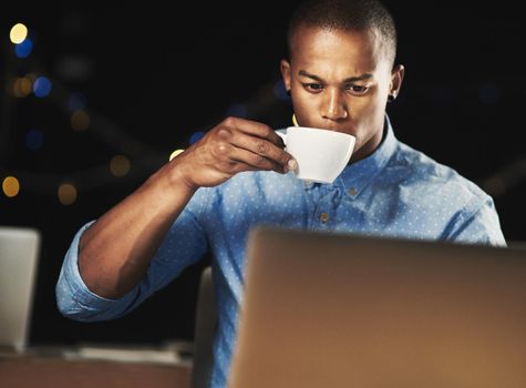 Just a little espresso to get him through the night. Shot of a young man drinking an espresso while working late night on his laptop.