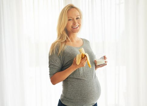 Healthy mommy, healthy baby. Portrait of a happy pregnant woman eating a banana and drinking a glass of milk at home.