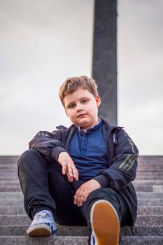 The boy is sitting on the steps in the open air against the background of skyscrapers and high-rise buildings. Journey. Lifestyle in the city. Center, streets.