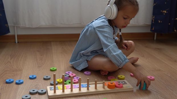 Happy Little Preschool Toothless Girl Playing With Colored Wooden Toy. Kids Learn To Count By Playing Teaches Numbers At Home. Child Listening To Music In Big White Headphones. Childhood, Education