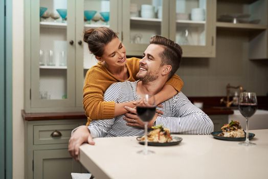 She sure knows the way to a mans heart. Shot of an affectionate young woman embracing her husband during supper in their kitchen at home.