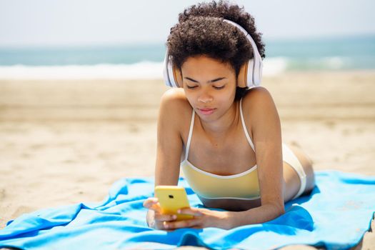 Young ethnic female tourist using smartphone and listening to music on beach