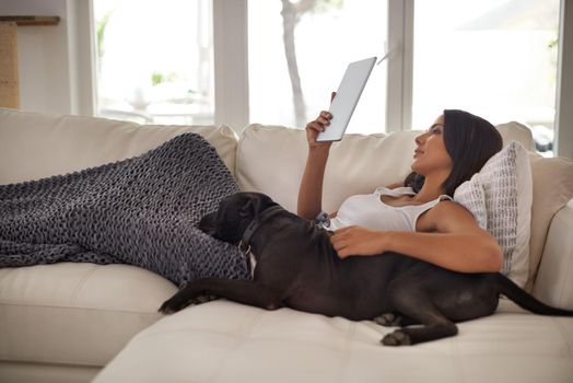 Staying connected from the comfort of my home. Shot of an attractive young woman relaxing on the sofa with her dog and using a digital tablet.