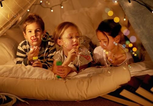 Camping in the bedroom. Shot of three young children in a blanket fort blowing bubbles.