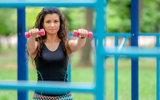 Pretty girl with dumbbells doing arm workout outdoors. Young woman exercising with sport fitness equipment at stadium in summertime