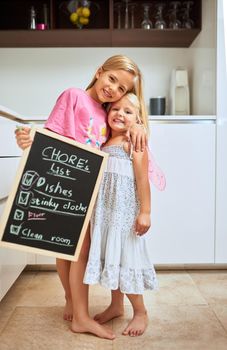 We did it together. Portrait of two little girl holding a chalkboard with a completed list of chores at home.