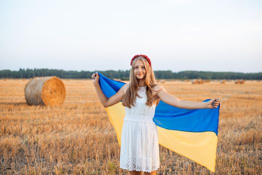 Young patriotic female with a flag of Ukraine in the harvested field of wheat with some bales of straw on the background