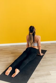 Side view of a beautiful young woman in sportswear practicing yoga on an exercise mat on a yellow background. Stretching the muscles of the back and legs, yoga