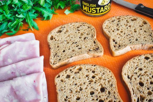 Slices of wholemeal brown bread laid out on a chopping board with ham, rocket and English mustard