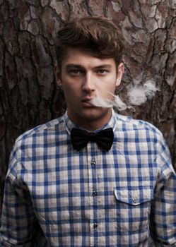 Getting some fresh air. Portrait of a young hipster smoking in front of a tree.