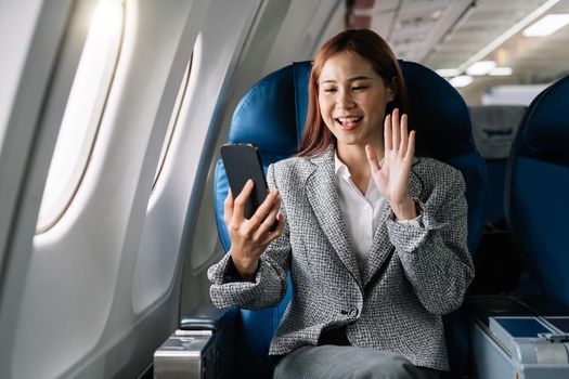 Smiling young asian businesswoman waving hand during video call on smartphone in plane