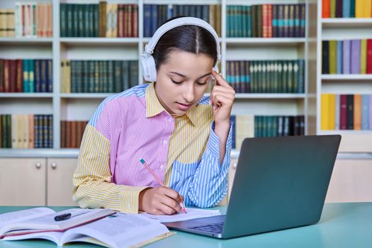 High school student girl in headphones studying in school library using books and laptop