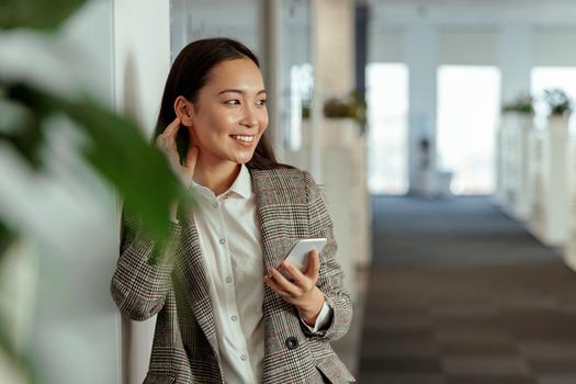Charming Asian woman with a smile standing holding mobile phone at the office and looking away