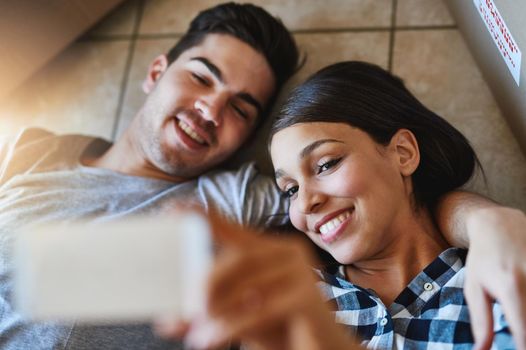 Crowning the moment with a selfie. Shot of a happy young couple taking a selfie while lying on the floor of their new home.
