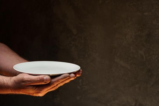 Male hands holding empty plate on dark background, lack of food, hunger concept.