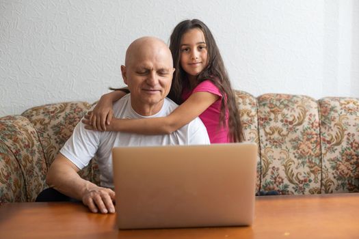 Happy little girl hugging smiling grandfather sitting on sofa with laptop.