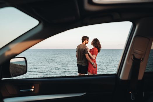 View Through Window of the Car Young Couple Standing on the Background of Sea, Man Hugging Curly Woman in Red Dress at Sunset