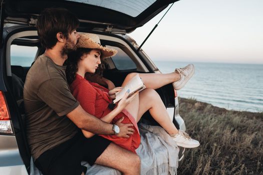 Man and Woman Sitting Inside Opened Trunk of Their SUV Car, Young Couple Enjoying Road Trip Along Sea at Sunset