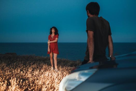 Man Sitting on the Hood of Car and Brunette Woman Dressed in Red Dress Standing in Front of Him Outdoors at Evening, Sea on the Background