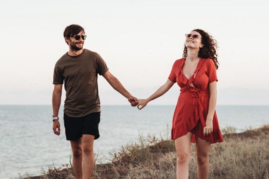 Cheerful Couple Walking Along the Sea, Man and Curly Woman in Red Dress Holding by Hands Enjoying Their Vacation