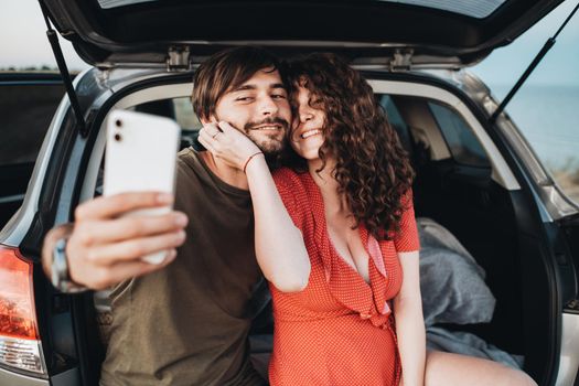 Happy Caucasian Couple Man and Curly Brunette Woman Making Selfie on Smartphone While Sitting in Trunk of the SUV Car
