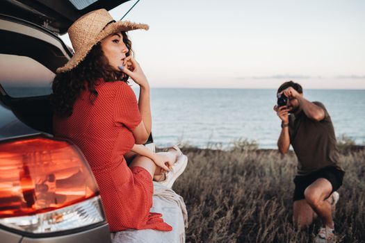 Man Photographing Woman in Red Dress and Hat That Sitting Inside Opened Trunk of the Car on the Background of Sea