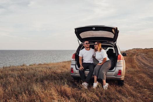 Happy Young Couple Sitting in Open Trunk of the SUV Car, Man and Woman Enjoying Their Road Trip Along Seaside