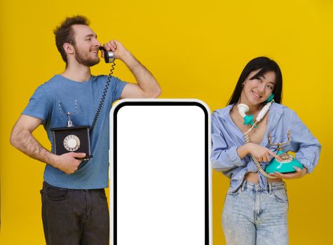 Handsome guy and asian girl talking on vintage phones leaned on huge smartphone or digital tablet with blank screen, happy smiling isolated on yellow background. Mock up, product placement