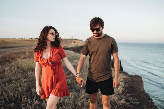 Cheerful Couple Walking Along the Sea, Man and Curly Woman in Red Dress Holding by Hands Enjoying Their Vacation