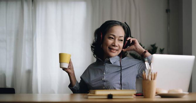 Happy senior asian woman wearing headphones listens songs on laptop in living room at cozy home
