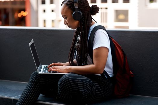 Get connected, get busy. Shot of an attractive young woman listening to music and using her laptop while relaxing outdoors.