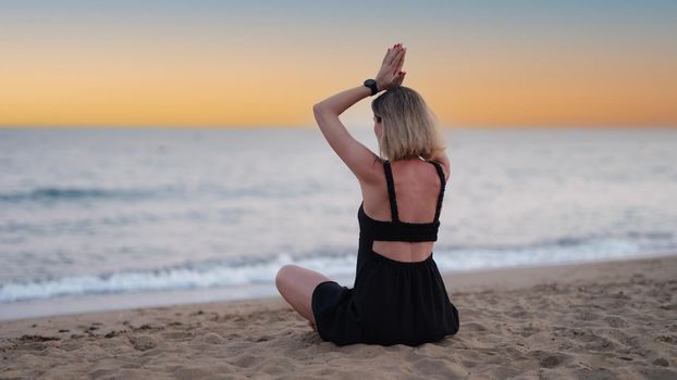 Woman doing yoga on seashore at sunset back view. Calmness and relax concept