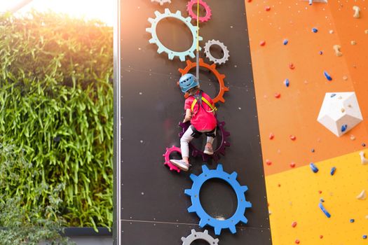 Child climbing on wall in amusement centre. Climbing training for children. Little girl in dressed climbing gear climb high. Extreme active leisure for kids.