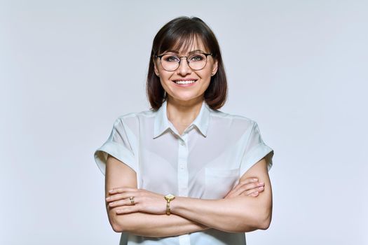 Portrait of positive confident middle aged woman looking at camera, light studio background. Smiling female in glasses, white shirt arms crossed. Business, professionalism, sales of goods and services